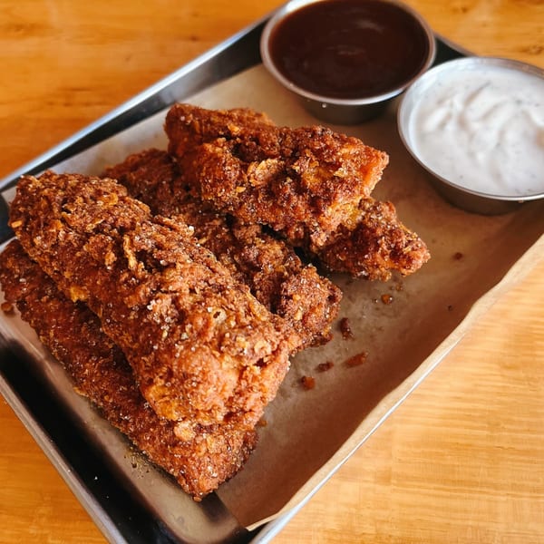 Crispy vegan chicken tenders with BBQ and ranch dipping sauces on a metal tray.